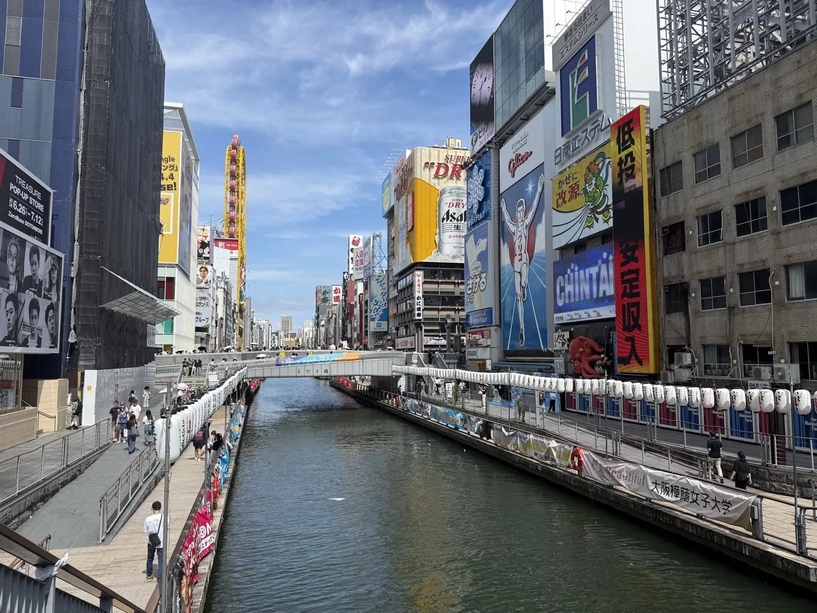 A wide panorama down the Dotonbori canal toward the Glico sign, neon signboards lining both banks and the riverwalk below