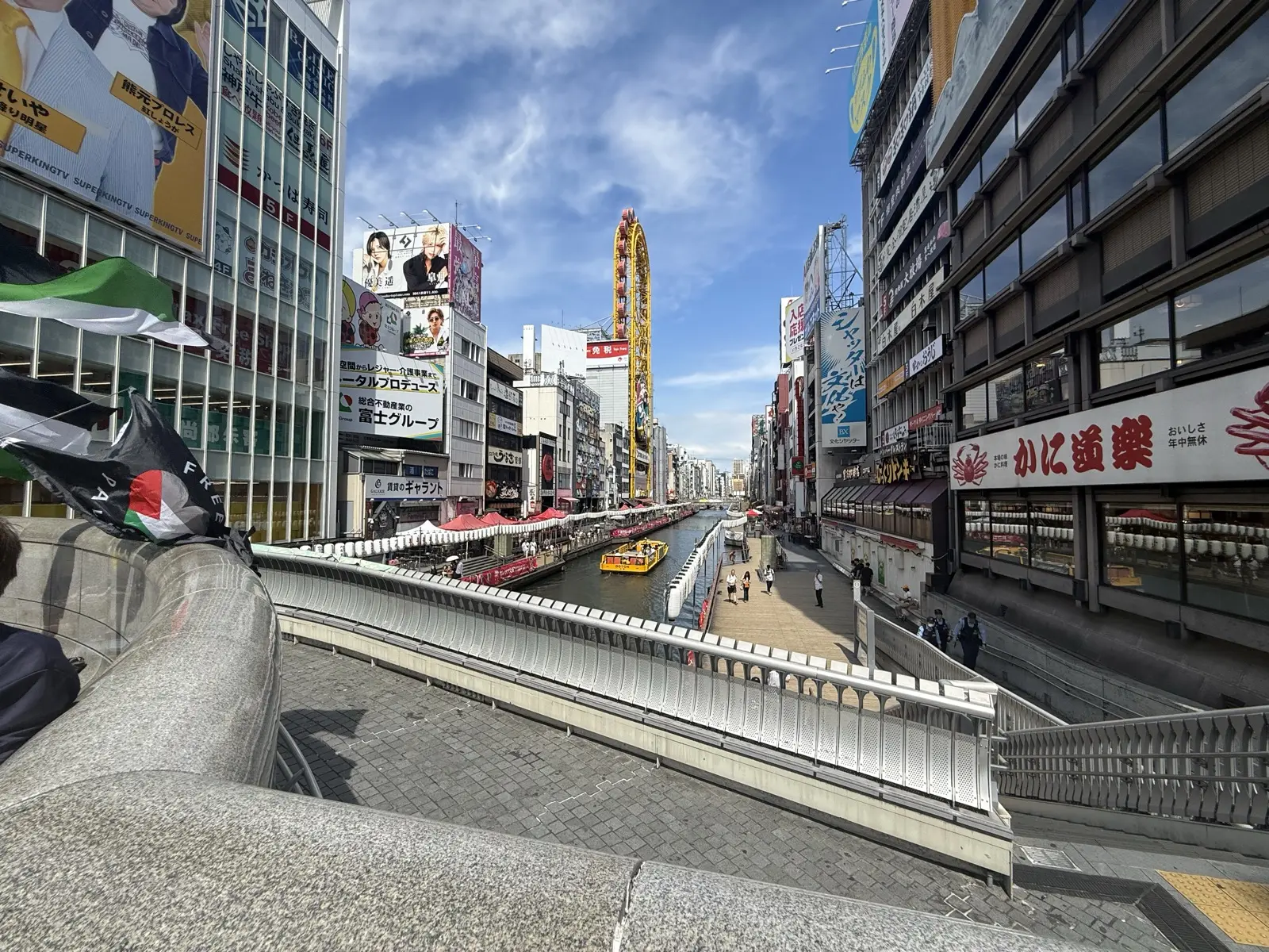 The Dotonbori canal with the Tombori Riverwalk, a sightseeing boat, the Ebisu Tower ferris wheel and a crab restaurant sign under a blue sky