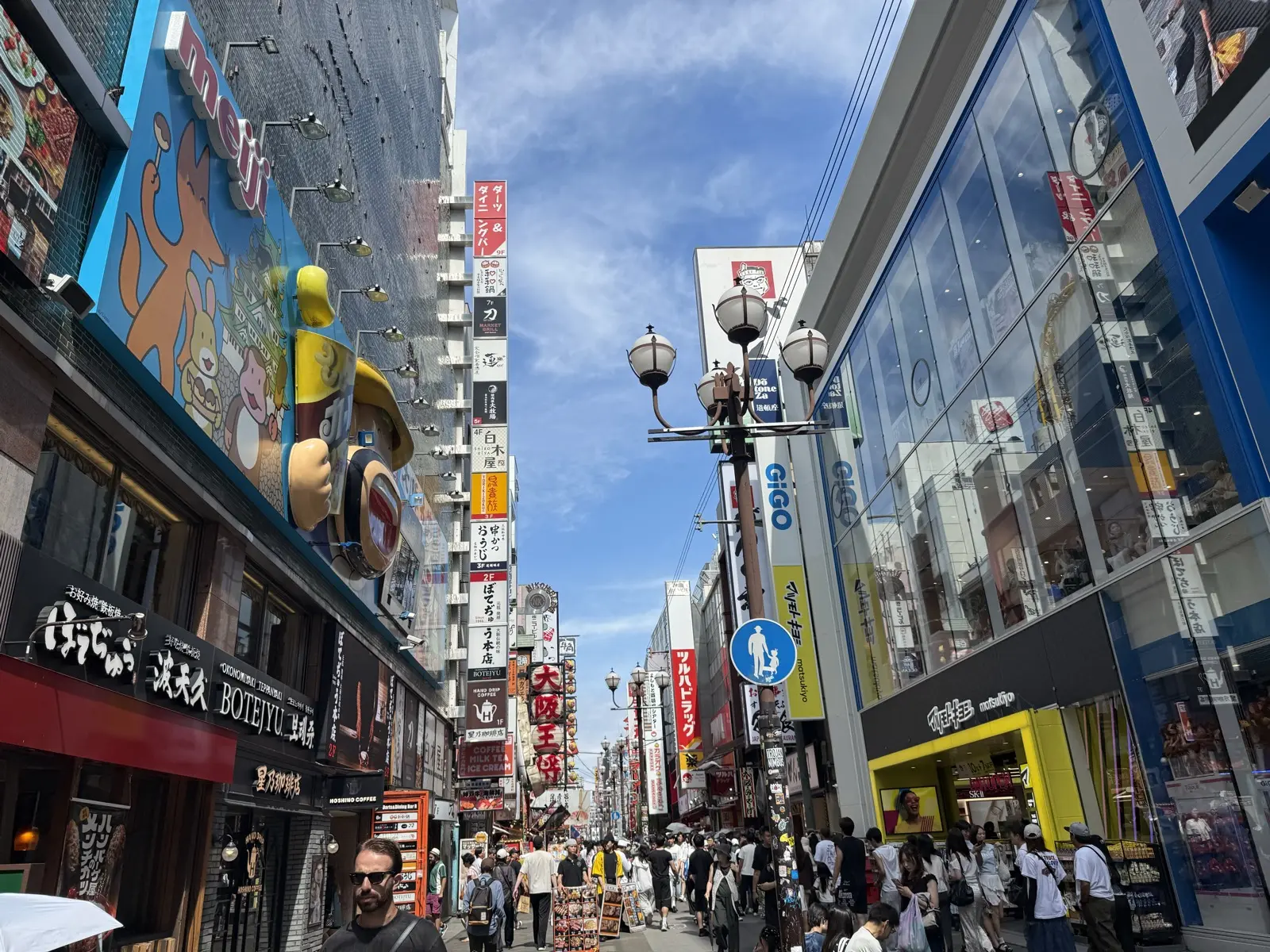 A busy food-and-shopping side street off Dotonbori in Osaka, with a Meiji 3D signboard, an okonomiyaki restaurant and game arcades