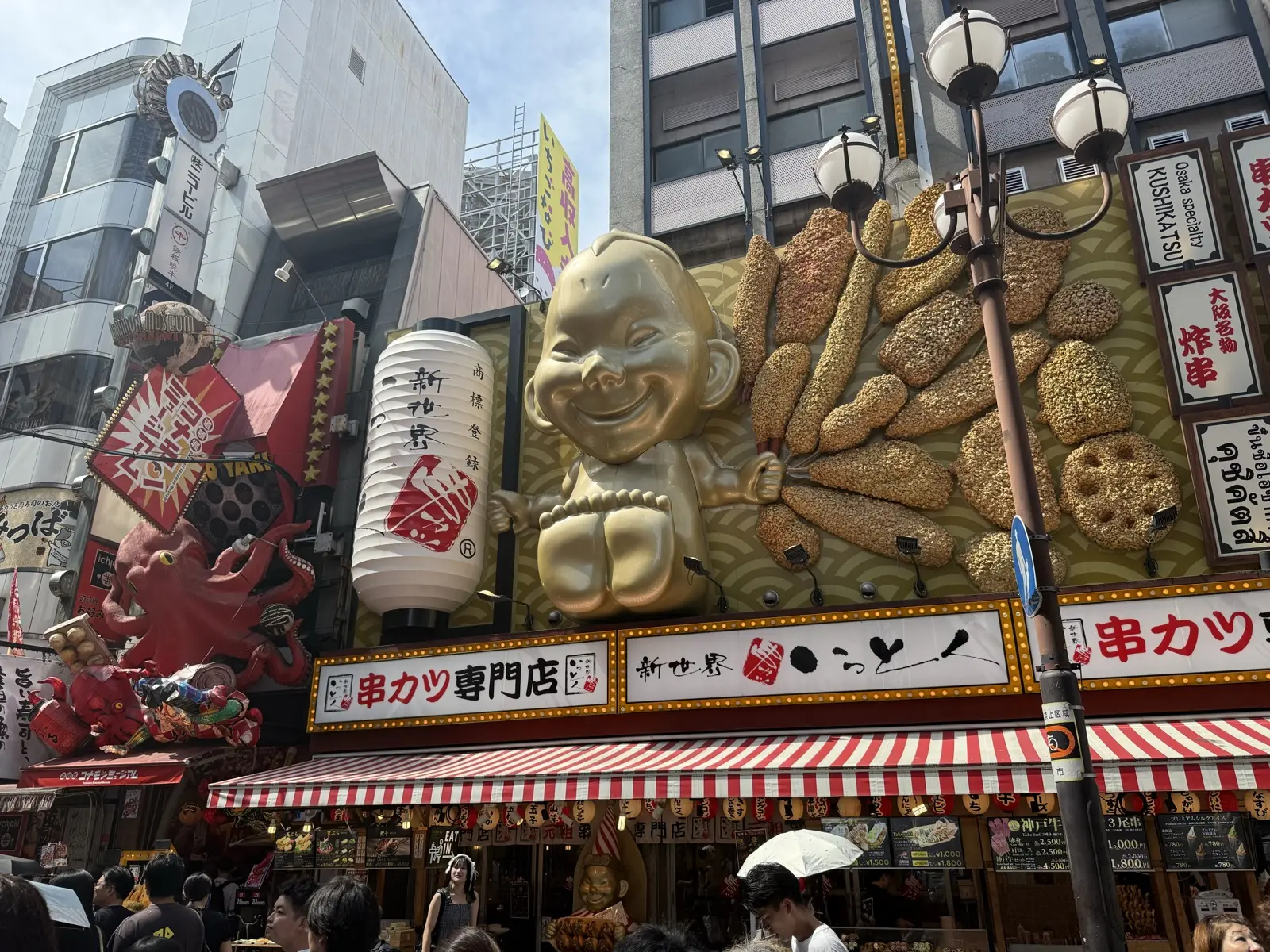 A giant gold Billiken good-luck figure perched over a kushikatsu shop in Osaka, surrounded by lanterns and skewer displays
