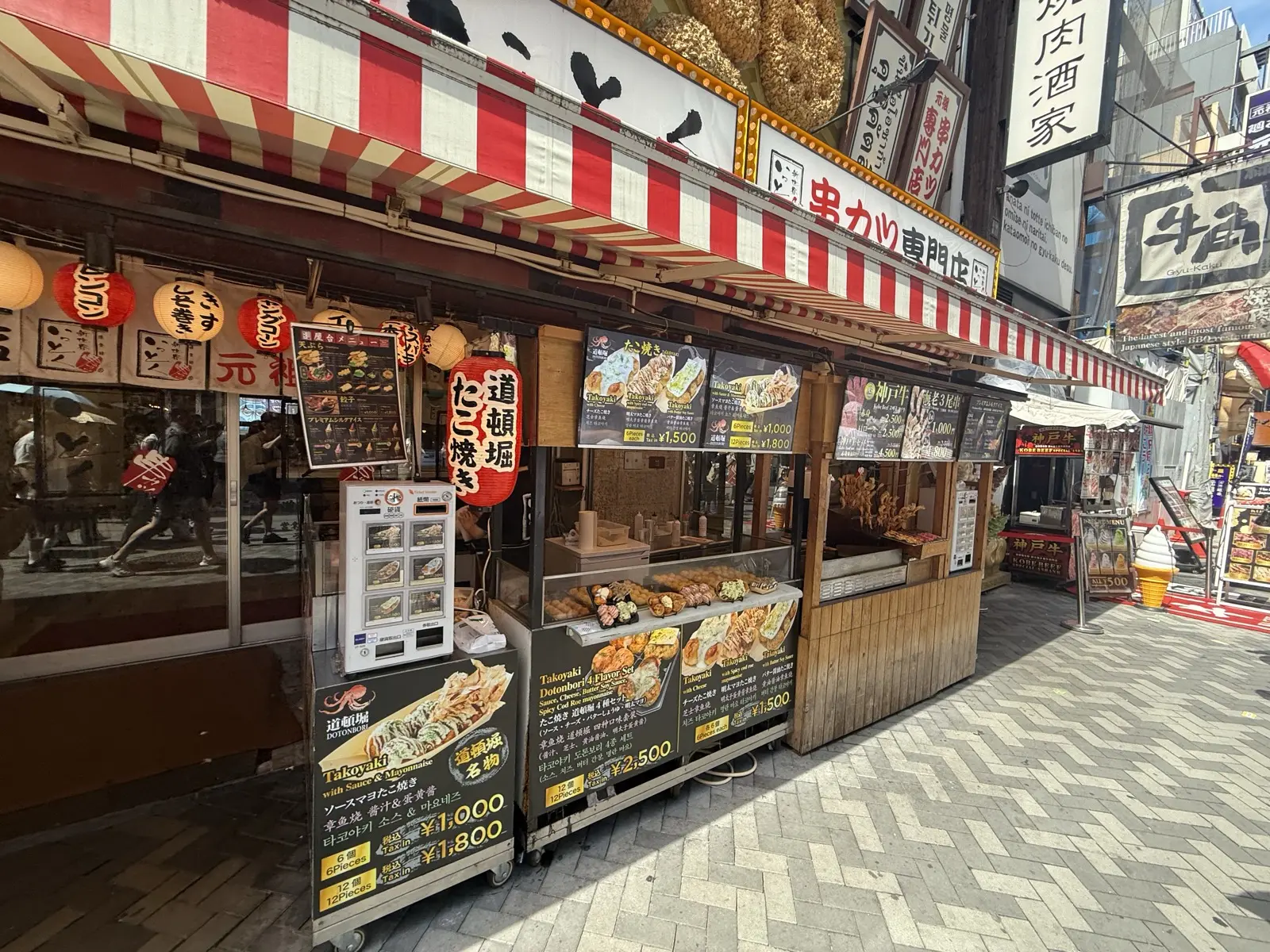 A Dotonbori street food stall selling takoyaki and kushikatsu, with photo menus and prices posted out front under a red-and-white awning
