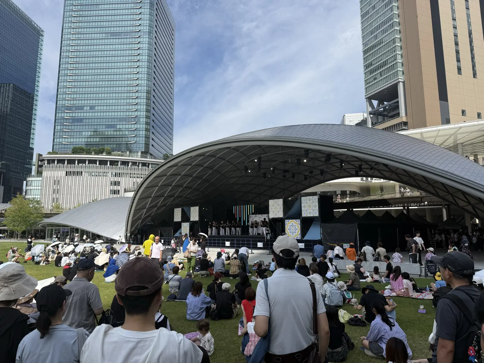 An open-air performance on the lawn at Grand Green Osaka (Umekita), with a crowd sitting on the grass in front of the curved-roof pavilion stage and skyscrapers behind