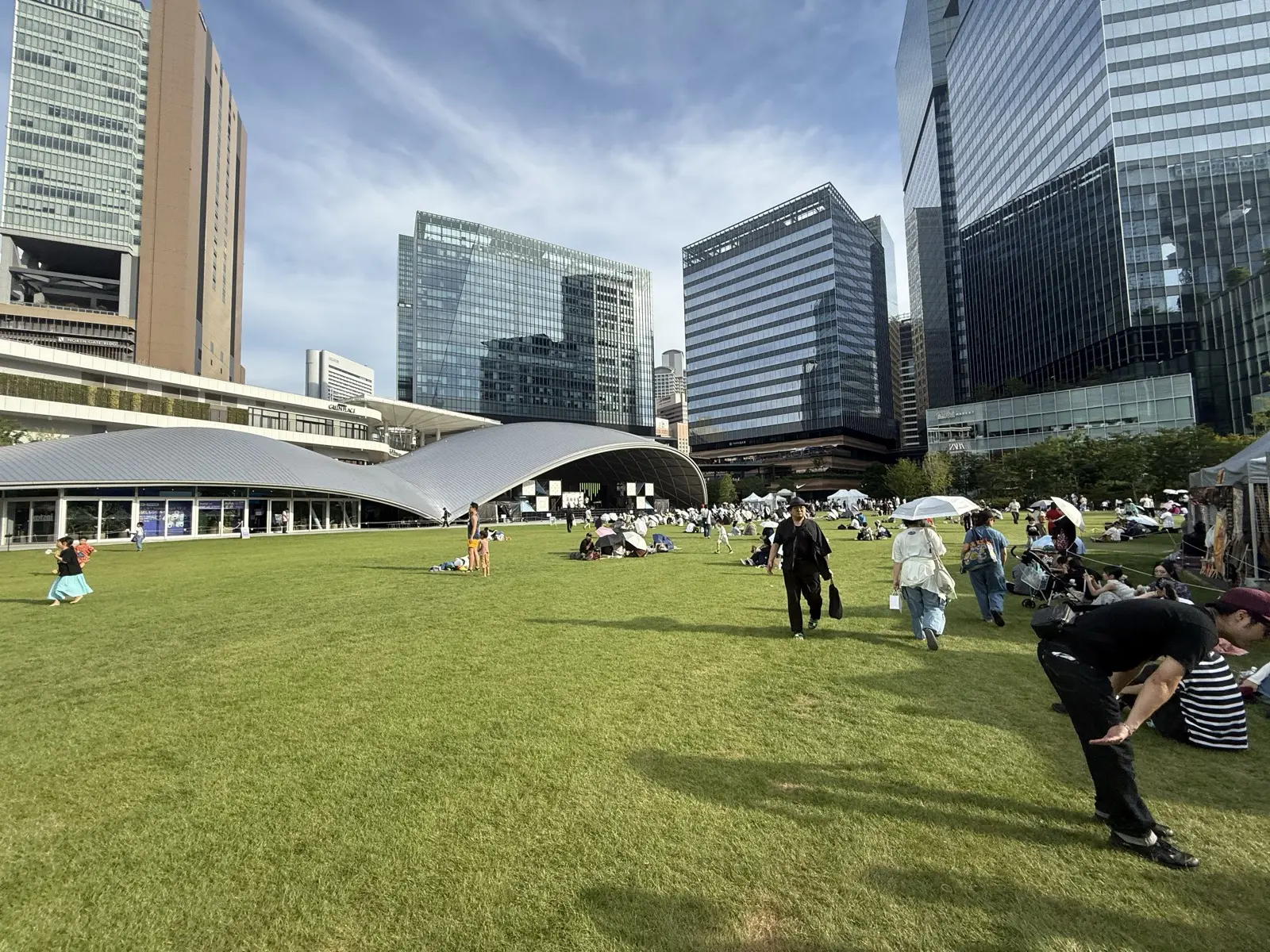The wide open lawn of Grand Green Osaka park with its curved-roof pavilion, people relaxing on the grass and market tents, framed by skyscrapers