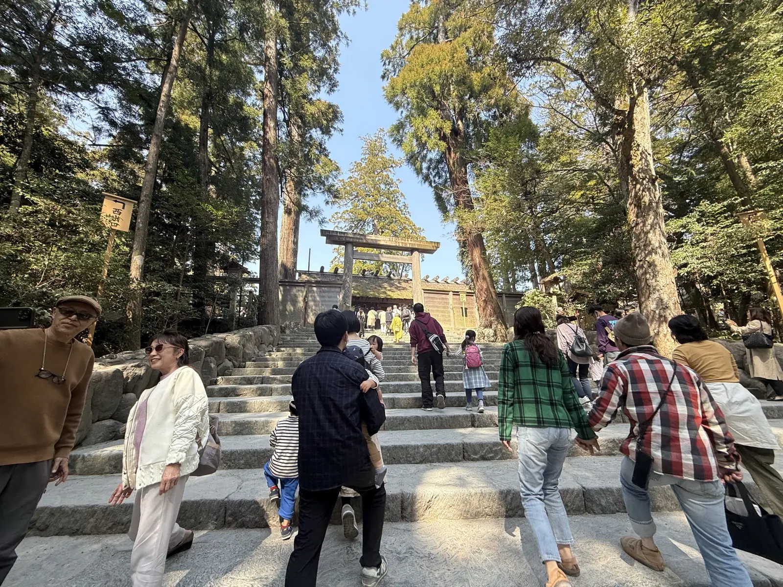 Stone steps rising through tall cedar trees to a plain wooden torii gate at the Naiku (Inner Shrine) of Ise Jingu, with visitors walking up