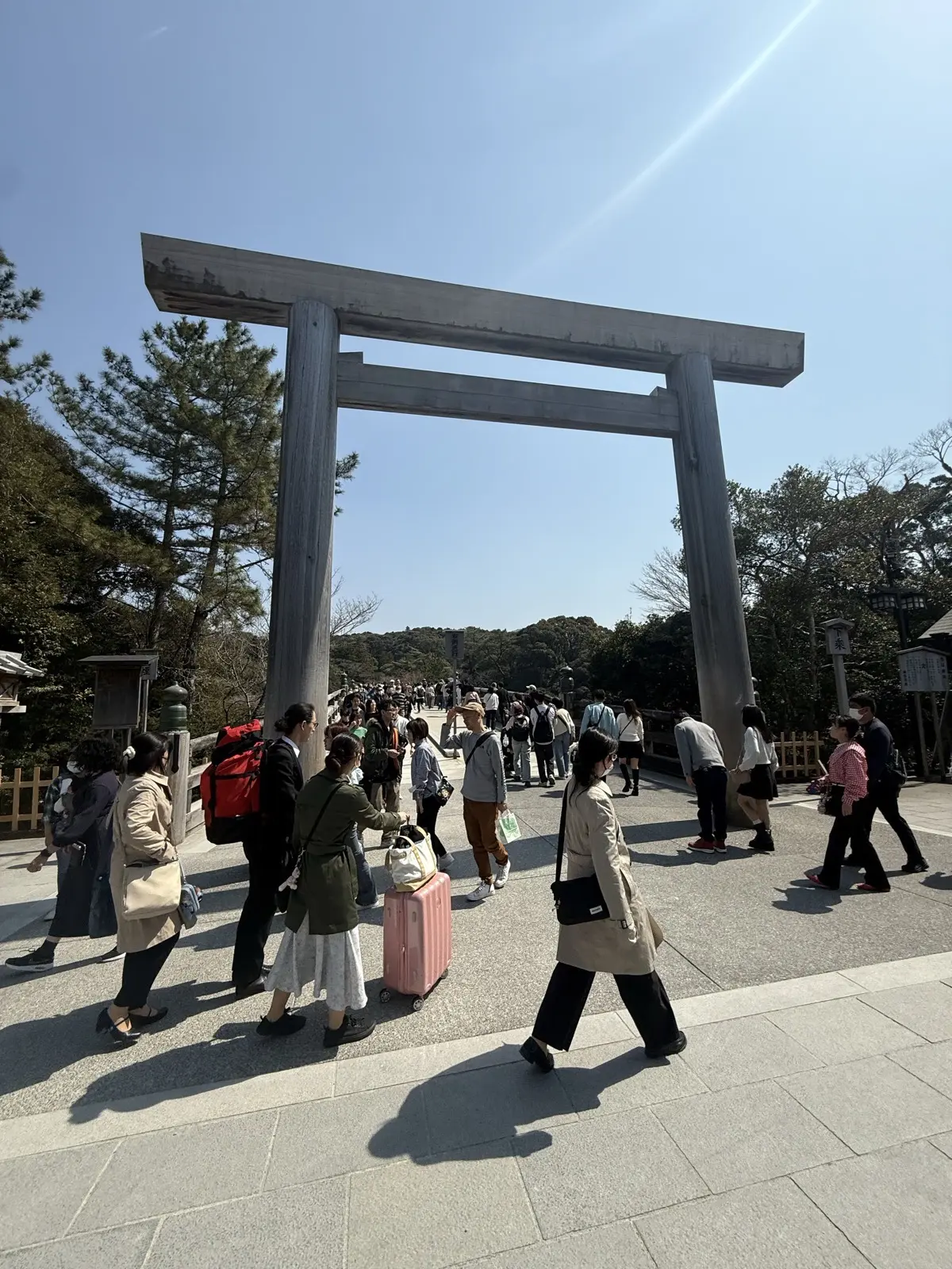 The large plain wooden torii gate at the entrance to the Naiku of Ise Jingu, with crowds of visitors and suitcases passing under it on a sunny day