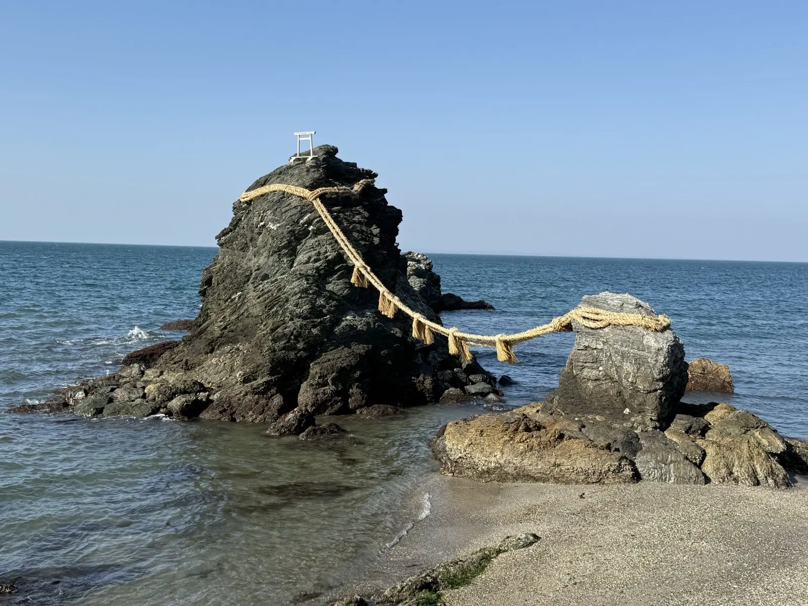 The Meoto Iwa Wedded Rocks at Futami near Ise — two sea rocks joined by a thick straw shimenawa rope, with a small torii on the larger rock, under a blue sky