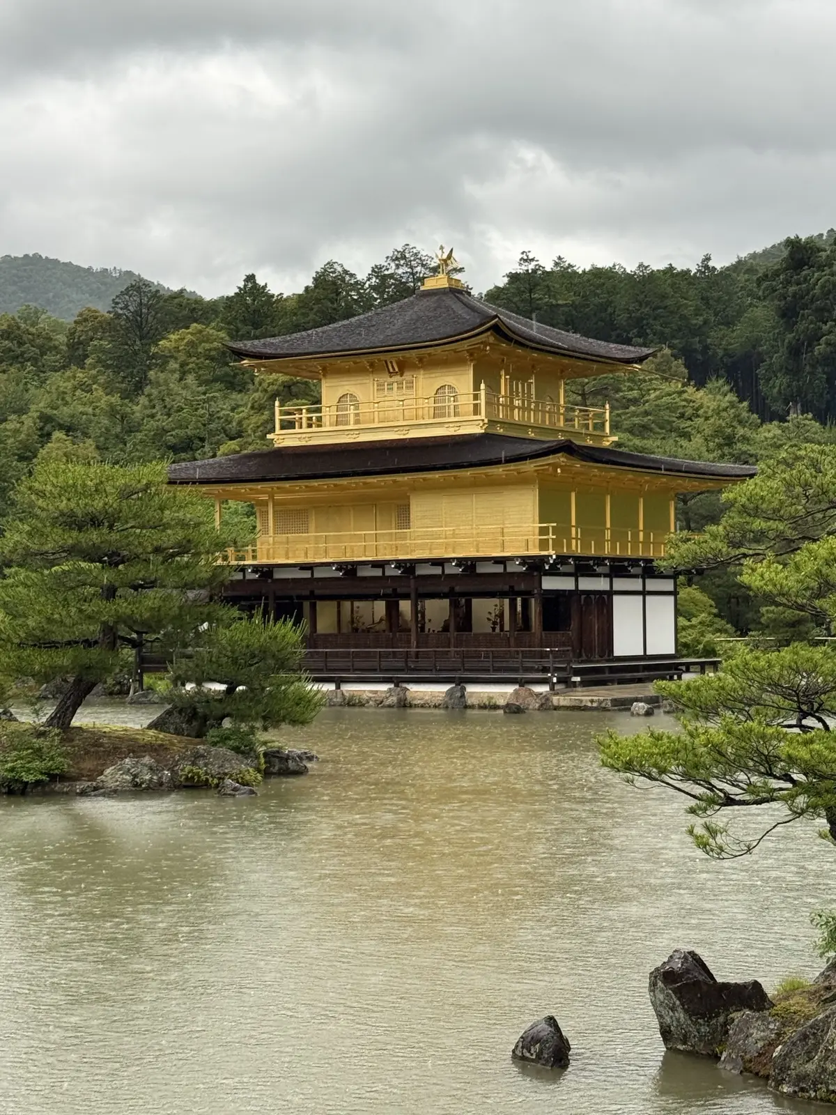 The Golden Pavilion reflected in the pond, framed by pines and hills — our own photo