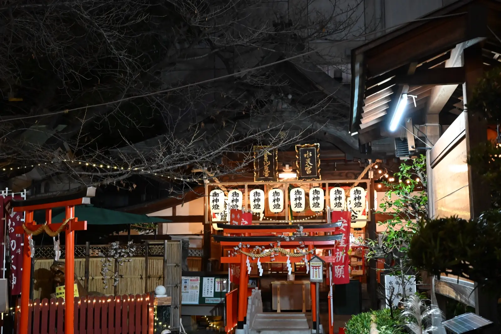Rows of lit paper lanterns reading Goshinto over a sub-shrine at Ohatsu Tenjin at night, with red torii and a safe-childbirth banner
