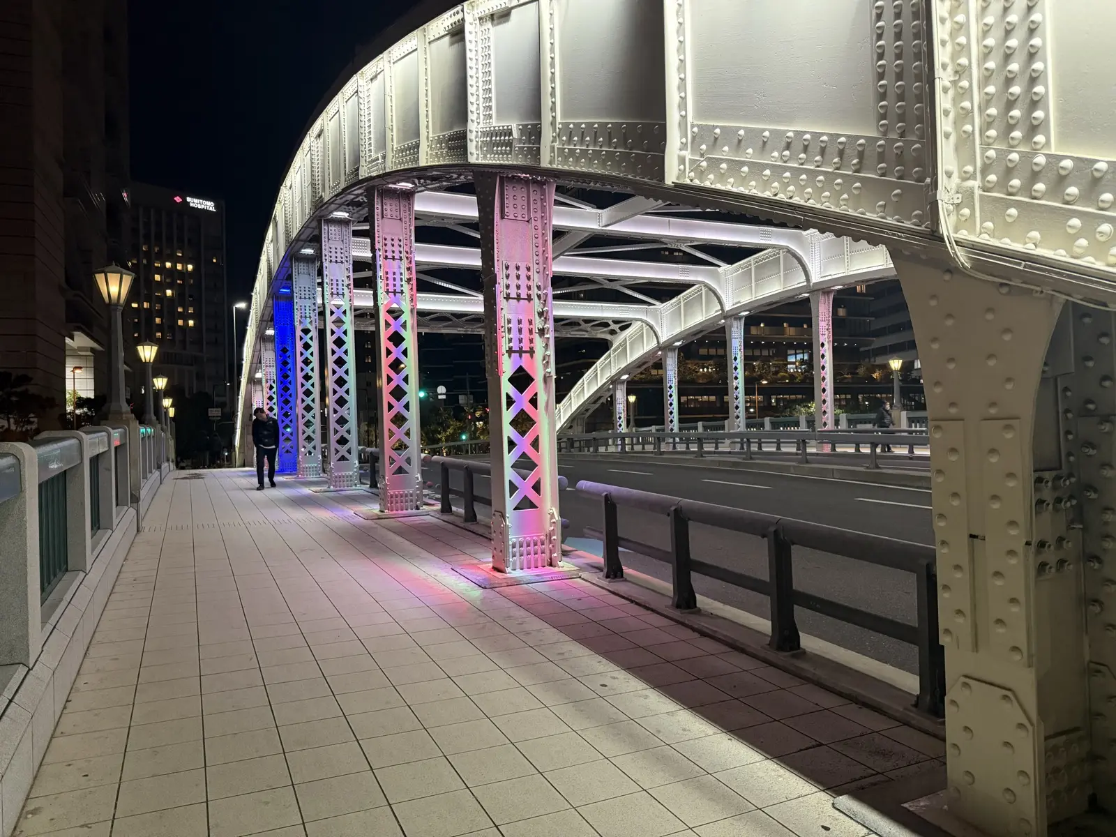 An old riveted-steel arch bridge on Nakanoshima lit up in pink and blue LED colours at night, with the pedestrian walkway