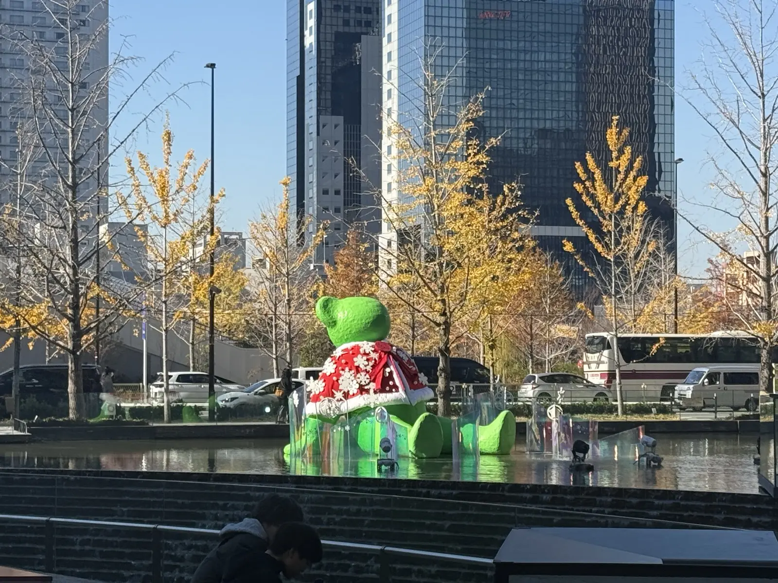 The green TED HYBER teddy bear sculpture by the water feature at Umekita Plaza in front of Osaka Station, with autumn ginkgo trees behind