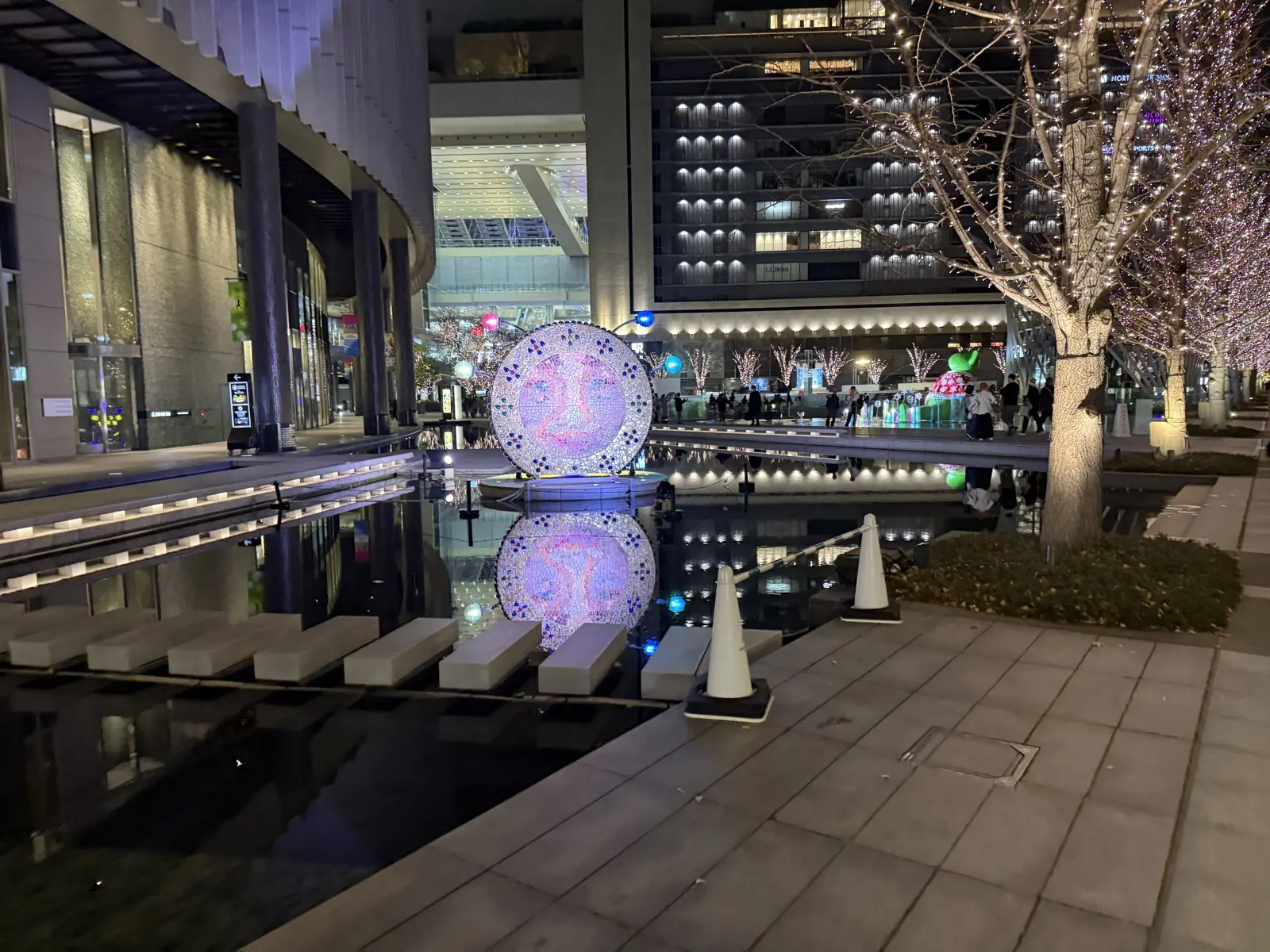 An illuminated circular light-art sphere reflected in a shallow pool at the Umekita plaza at night, with lit trees behind