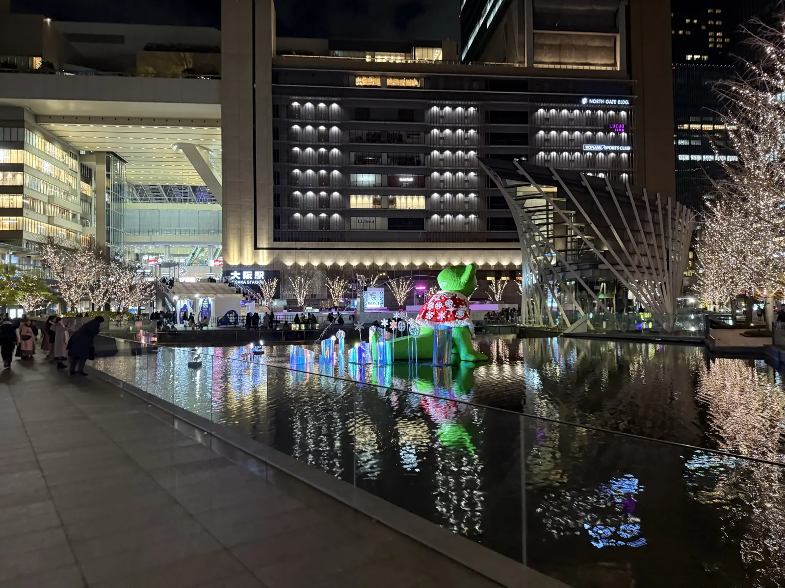 The water plaza outside Osaka Station at night, with the TED HYBER green teddy bear sculpture, illuminated trees and the North Gate building all reflected in the shallow water