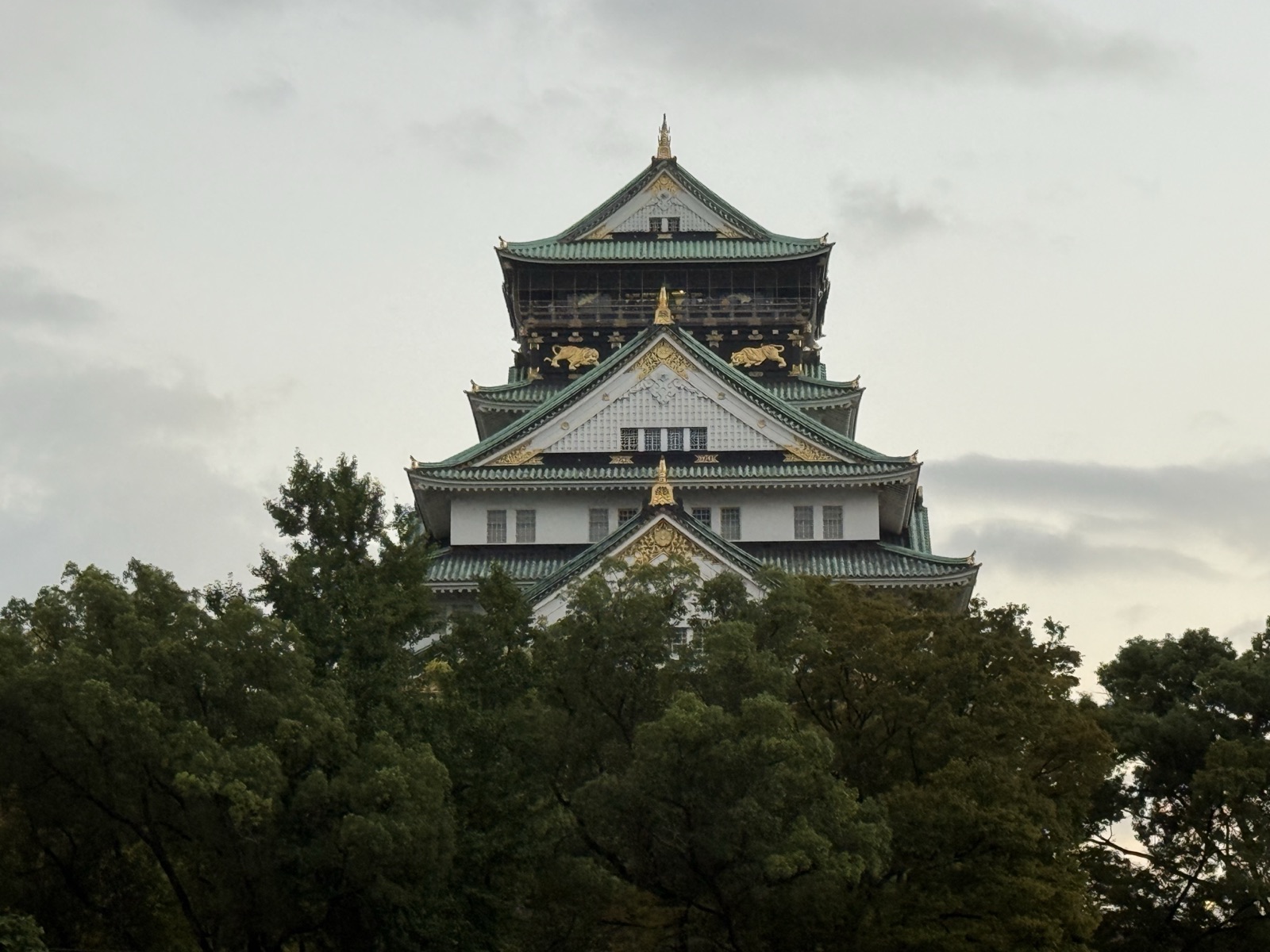 Close-up of Osaka Castle's main tower showing green roofs and gold tiger and crane ornaments above the trees